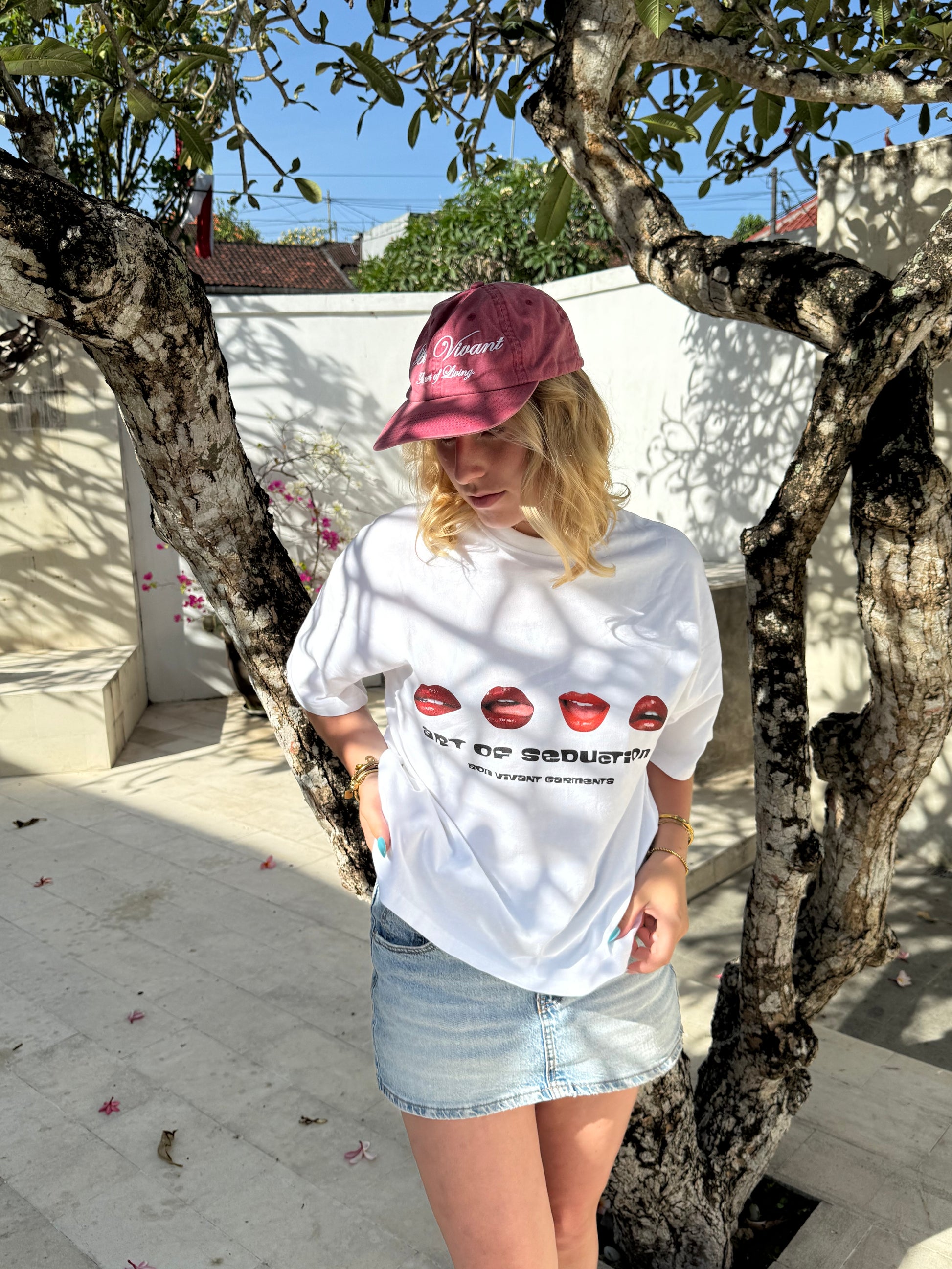 Person wearing a white hoodie with red graphics and a pink cap, leaning against a tree.