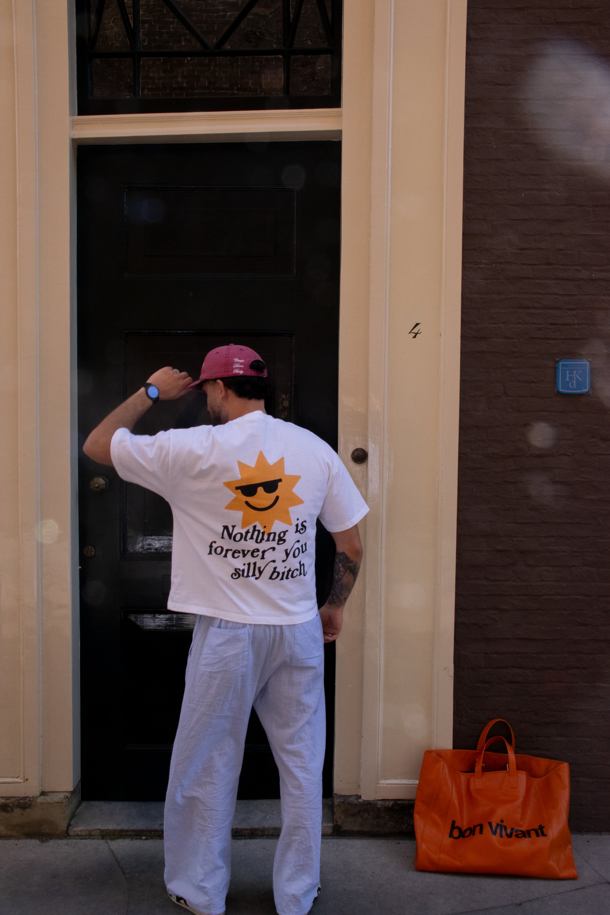 Person wearing a white t-shirt with a sun graphic and text, standing next to an orange bag on a sidewalk.